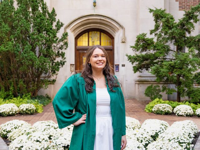 Image of Honors College student Brandi Stover standing with a hand on one and smiling at the camera. Brandi is wearing a green graduation gown and white dress. White flowers, a red brick path, green shrubs, and the tan stone of MSU's Beaumont Tower are in the background.