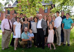 Image of Gary Seevers and spouse Eiko stand on the grass in front of a red brick building on Michigan State University's campus. There are lush tree branches above them, and they are surrounded by a multitude of multi-generational family members. Everyone is in business casual and business formal attire and smiling.