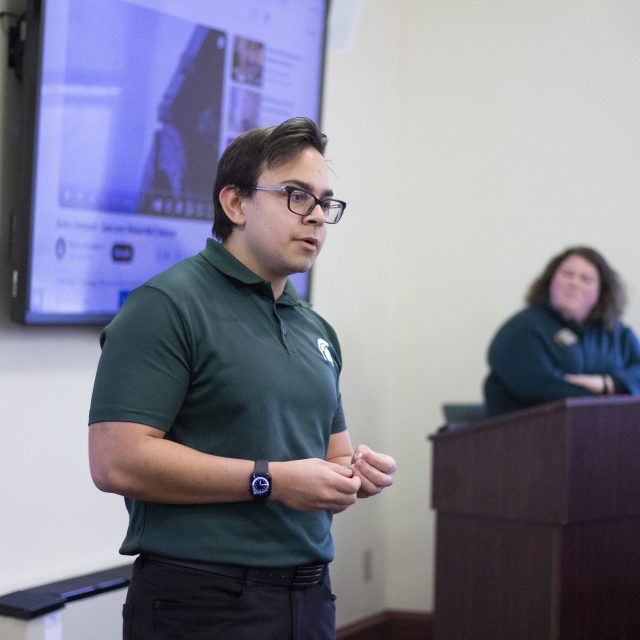 Image of Honors College student Brennan Haugen presenting at the 2024 Honors Giving Back Awards. Haugen is in a green polo shirt and black dress pants, standing in front of a large presentation screen.