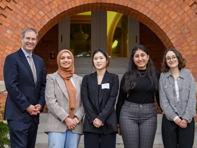 MSU Honors College Dean Chris Long stands with four students in front of the red brick arched entrance of Eustace-Cole Hall. Most everyone is smiling, and all of them are wearing business professional attire.