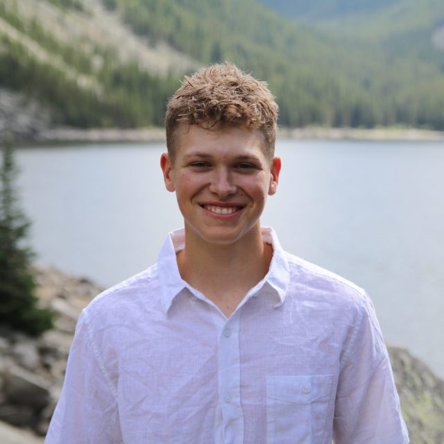Blaise Maliskey profile photo wearing a white button-up shirt and standing in front of a lake and green mountains.