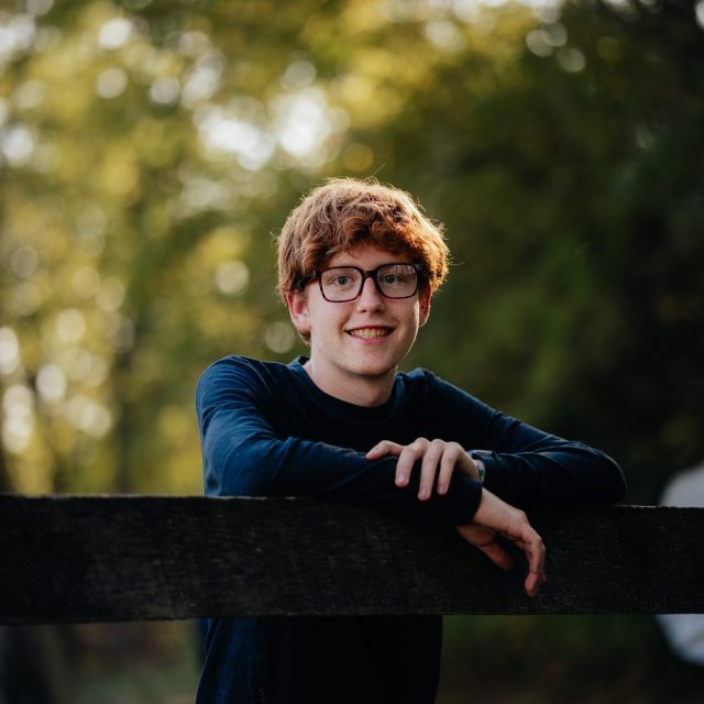 Connor Mason profile photo leaning on a railing with greenery in the background.