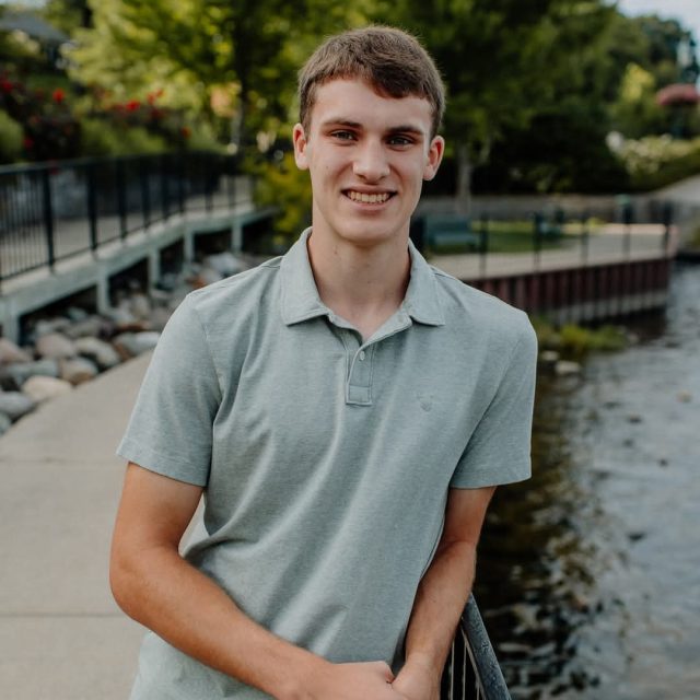 David Stout profile photo leaning on a bridge railing with trees and water in the background.