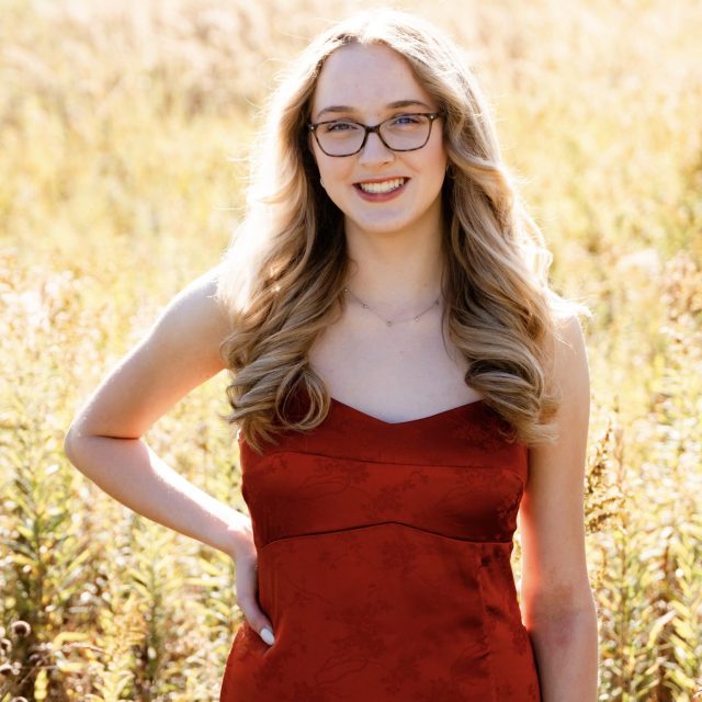 Emily Sklenicka profile photo wearing a red dress and standing in a field of grass.