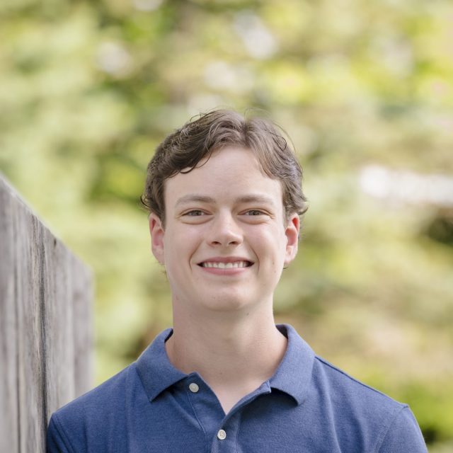 Nathan Romito profile photo wearing a blue shirt with greenery in the background.