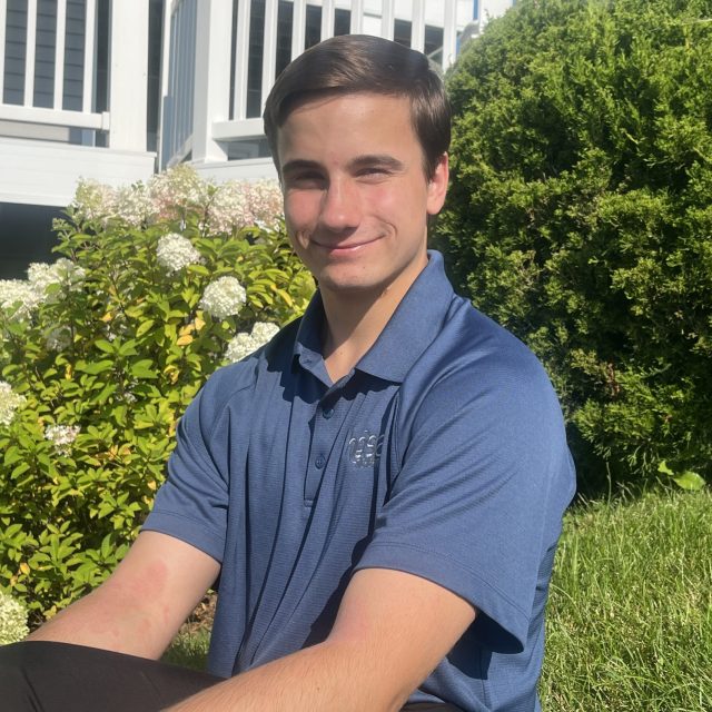 Peter Sanin profile photo wearing a blue shirt with greenery and a white fence in the background.