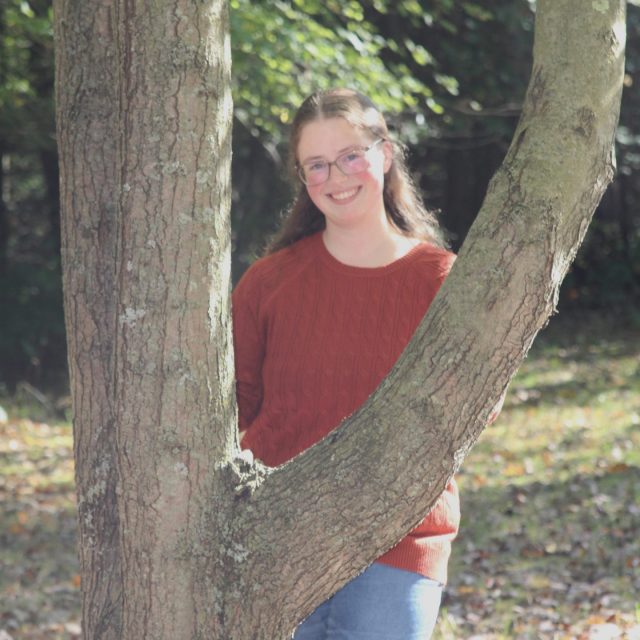 Samantha Wagner profile photo wearing jeans and a red top, standing by a tree with greenery in the background.