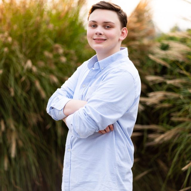 William Daily profile photo wearing a light blue button up shirt and standing in front of tall grasses.