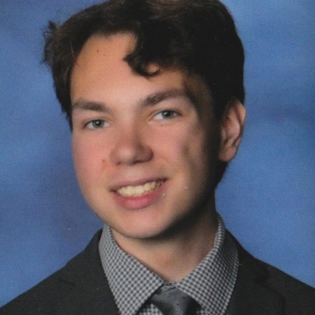 William Ford profile photo wearing a dark gray suit and gray neck tie, in front of a neutral blue background.