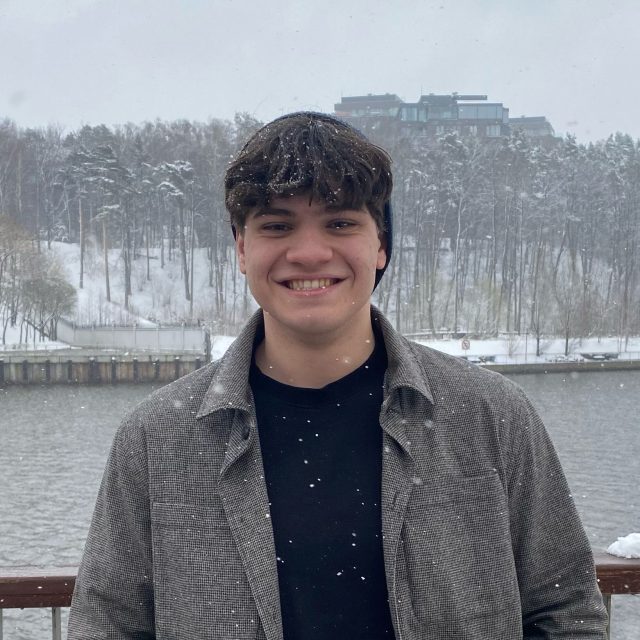 Aleksandr Skorkin profile photo wearing a black shirt and gray coat standing in front of a snowy waterfront.