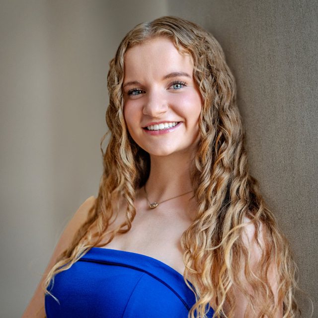Brianna Deeble profile photo wearing a blue top and leaning against a wall in the beige background.
