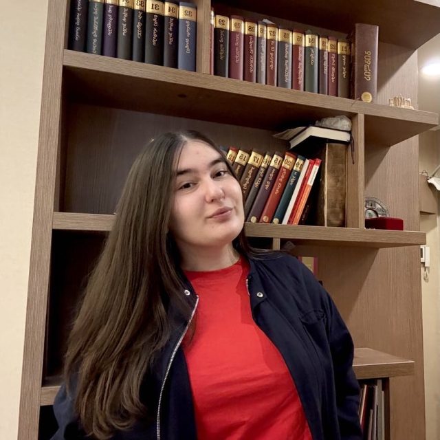 Ana Toradze profile photo wearing a red shirt and dark jacket and standing in front of a brown bookcase full of books.