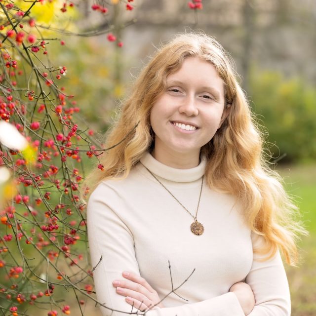 Anja Beck profile photo wearing a white turtleneck and standing by a bush of red berries.