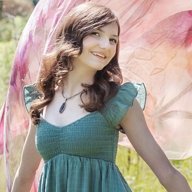 Anneke Anglin profile photo wearing a green dres and holding a large pink fabric that blows in the background.