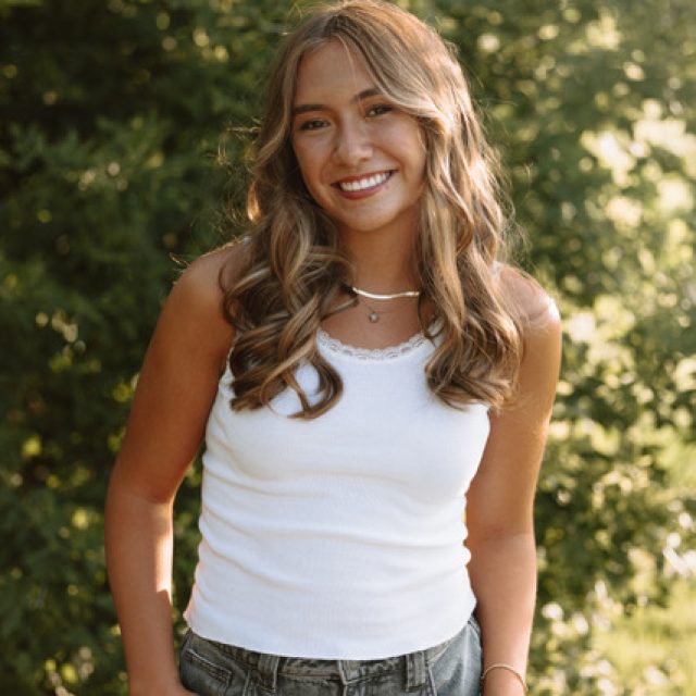 Ava Crozier profile photo wearing a white tank top and standing in front of greenery.