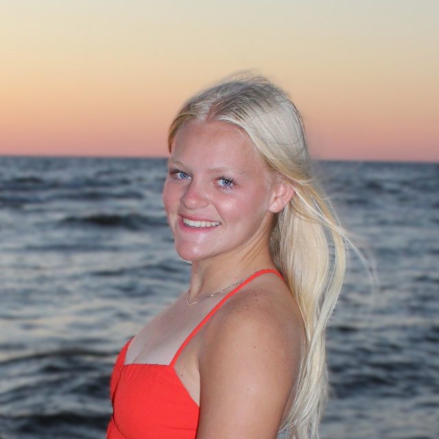 Ava Freye profile photo wearing a red tank top with a waterfront in the background during sunset.