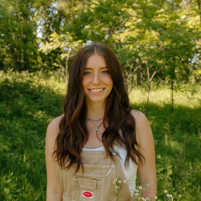 Avery Riehl profile photo wearing beige overalls with greenery in the background.