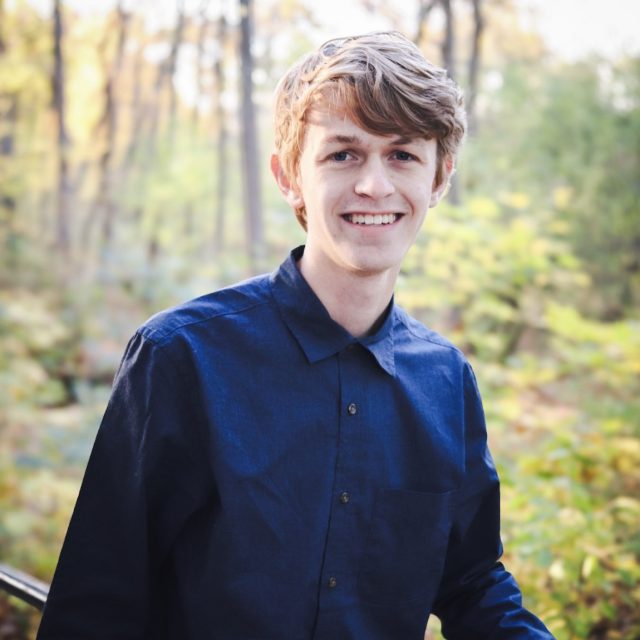 Harrison Cornell profile photo wearing a blue button-up shirt with a forest in the background.