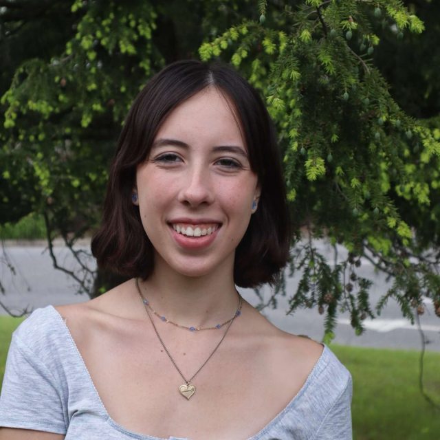 Miriam Nell profile photo wearing a white top with greenery in the background.