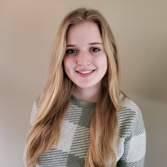 Nora Thornber profile photo wearing a gray and white plaid sweater and standing in front of a neutral white background.
