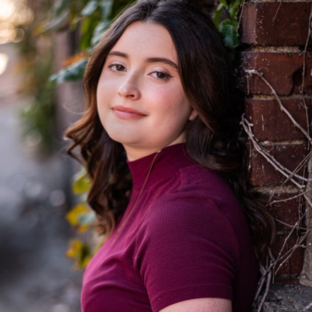 Rylee Gehrlein profile photo wearing a maroon top and leaning against a brick wall covered in ivy.