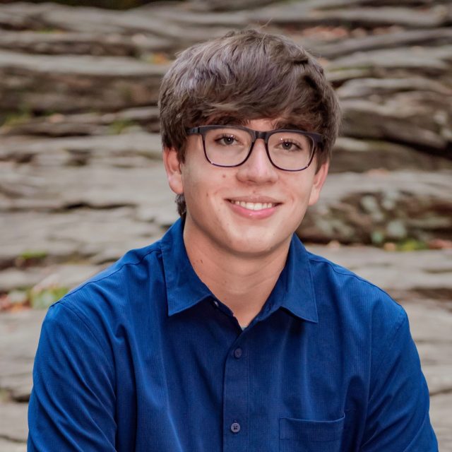 Sean Burk profile photo wearing a blue button-up shirt with gray rocks in the background.