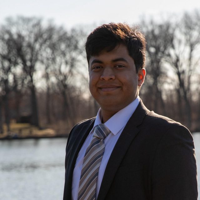 Tejas Agrawal profile photo wearing a black suit with a striped neck tie. There is a waterfront lined with trees in the background.