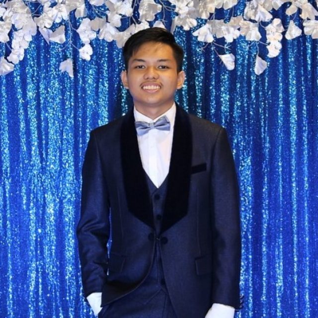 Yesi Doctolero profile photo wearing a navy blue suit and a light blue bowtie in front of a glittery blue background.