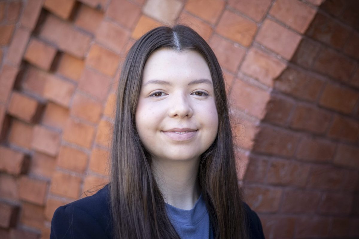 Image of Eleanor Pugh smiling. Pugh is in a dark blazer and blue shirt, standing against a red brick background.