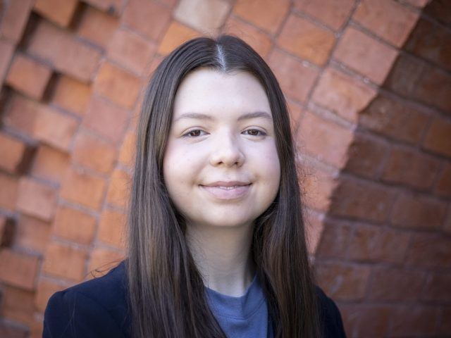 Image of Eleanor Pugh smiling. Pugh is in a dark blazer and blue shirt, standing against a red brick background.