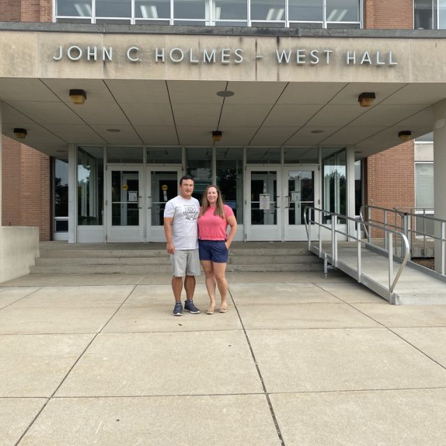 Image of Michigan State University alumni Michelle and Randy Kimple standing in front of a residence hall in shorts and t-shirts. The words on the entryway above them read: JOHN C. HOLMES - WEST HALL.
