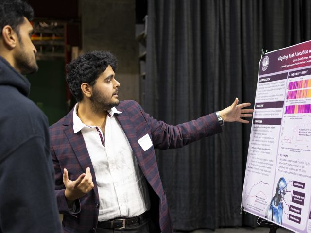 Image of Dhruv Kekin Toprani in a plaid maroon and navy suit jacket gesturing to a large research poster.