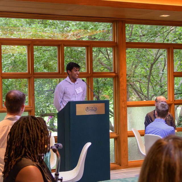 Image of Honors College student Aaron Philip standing and smiling at a green and gold podium. Donors are attentively seated at tables around Philip.