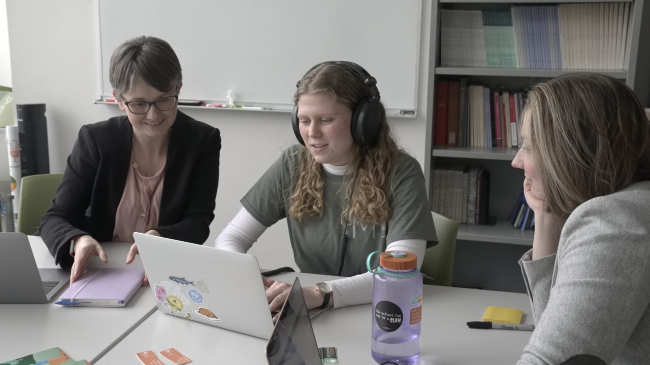 Caroline Zackerman in a classroom setting wearing headphones on a laptop and alongside two mentors.
