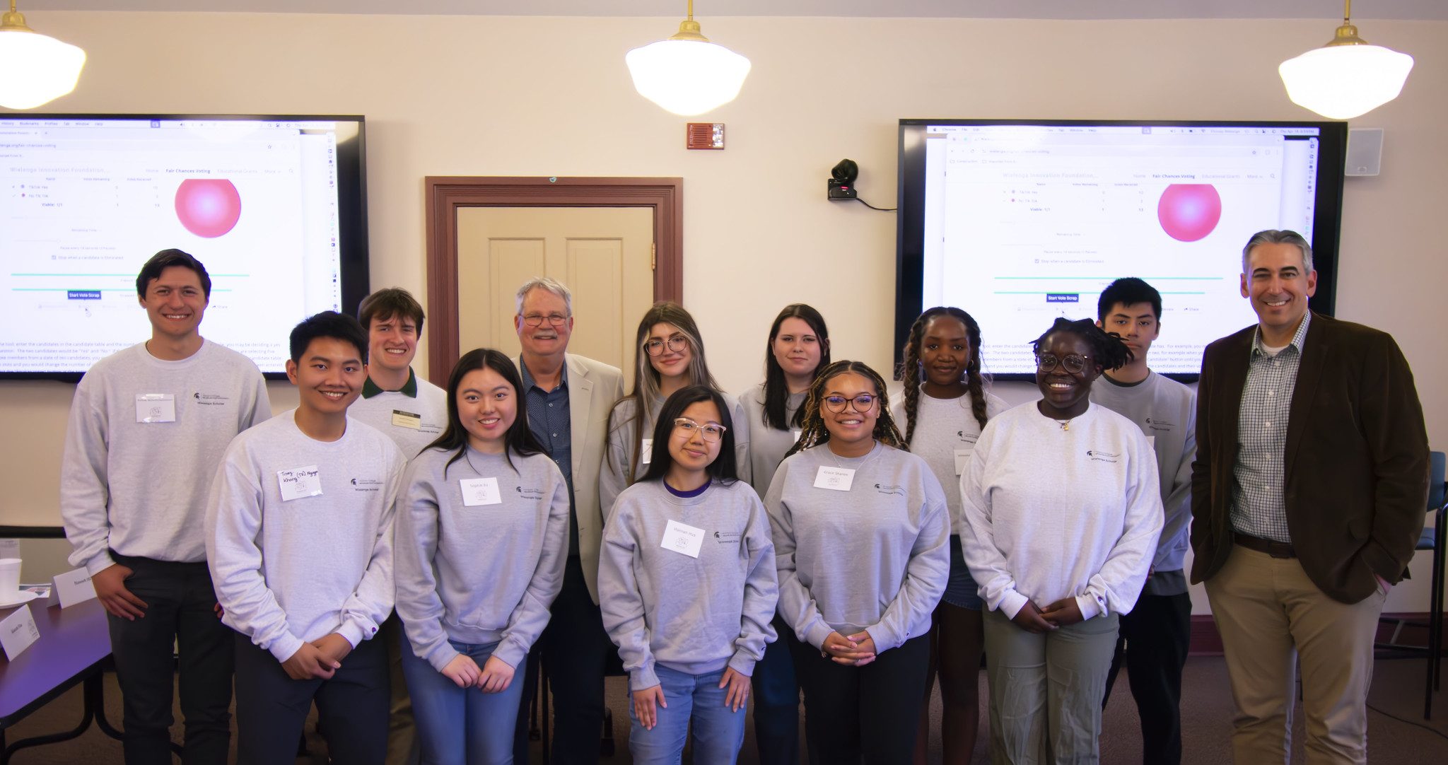 Image of a group of smiling of Wielenga Research Scholars in grey sweatshirts. They are standing in a room with Thomas Wielenga and Honors College Assistant Dean Justin Micomonaco, both in business attire. Two large presentation screens are in the background.
