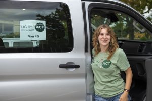 Image of Maggie Broderick in a green HC IMPACT mentor t-shirt, stepping out of the passenger-side of a grey van and smiling. There is a sign in the van window that reads: HC IMPACT Van #5.