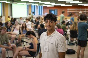 Portrait of Anas Shaaban in a white polo and name tag, smiling in a classroom with HC IMPACT students in the background. 