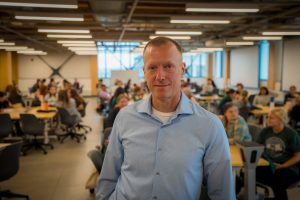Portrait of Neil Steinkamp wearing a blue button-up and smiling at the camera. In the background is a class full of IMPACT participants. 