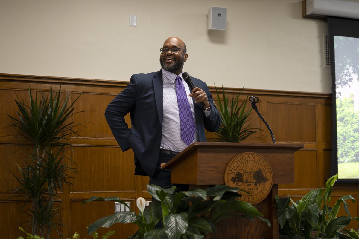 Image of Honors College Interim Dean Glenn Chambers standing at a wooden podium inside Campbell Hall holding a microphone. Chambers is wearing a dark suit jacket, purple tie, and light purple collared shirt and smililng.