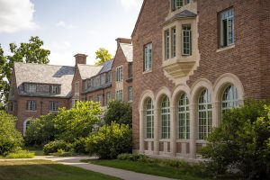 Image of the front of Michigan State University's Campbell Hall from a side angle. The brown brick building has stone archways, teal windows, and a lush green lawn and shrubbery.