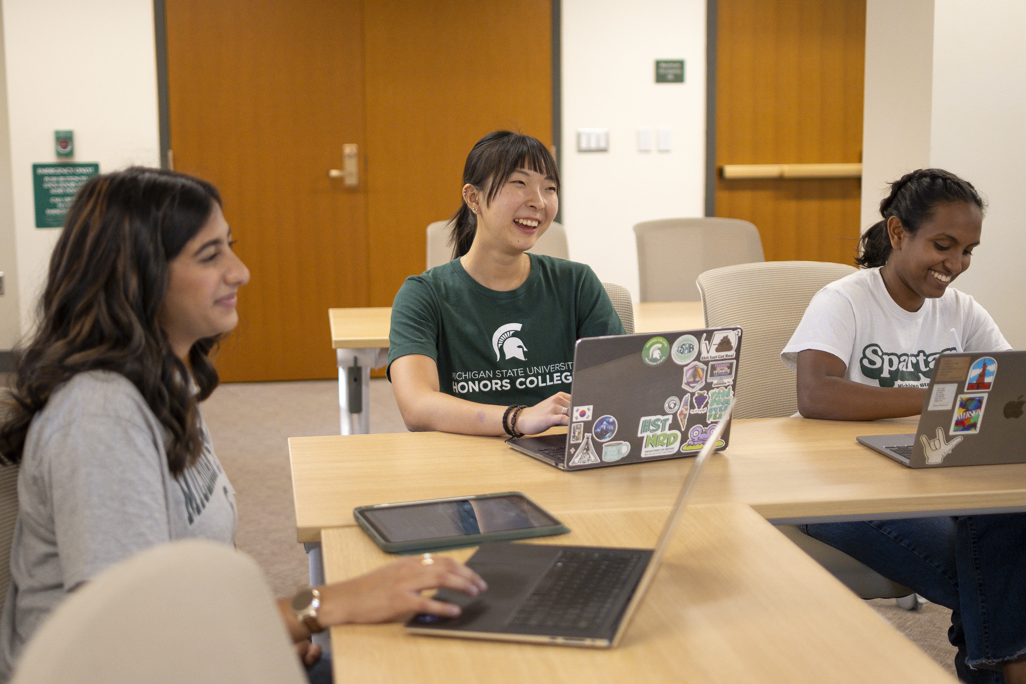 Image of Michigan State University Honors College students Anusha Pandya, Kara Hwang, and Ann Joseph sitting at a table in a classroom at Campbell Hall. They are in various MSU t-shirts, interacting with their laptops and smiling.