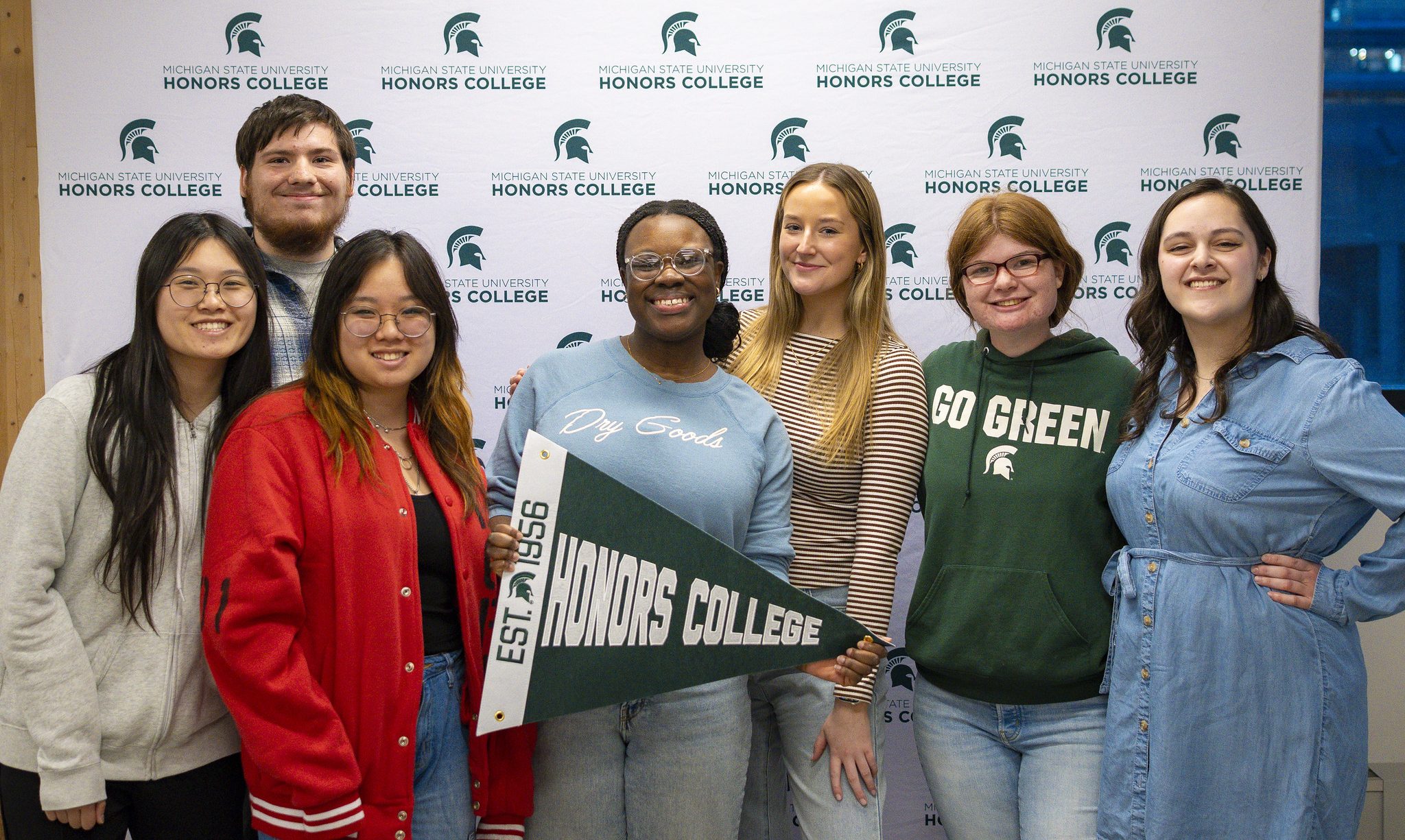 Image of Michigan State University student leaders for the Academic Scholars Program Advisory Council. The group of seven students is standing in front of a white backdrop with a pattern of repeating dark green MSU Honors College logos. The students are smiling at the camera in casual apparel. One is holding a dark green and white Honors College pennant.