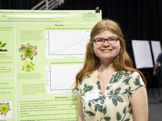 Image of Michigan State University student Basia Love wearing a cream-colored top with dark green floral patterns. Love is smiling at the camera, wearing a name tag, and staging in front of a large green poster with plant images on it at the University Undergraduate Research and Arts Forum.