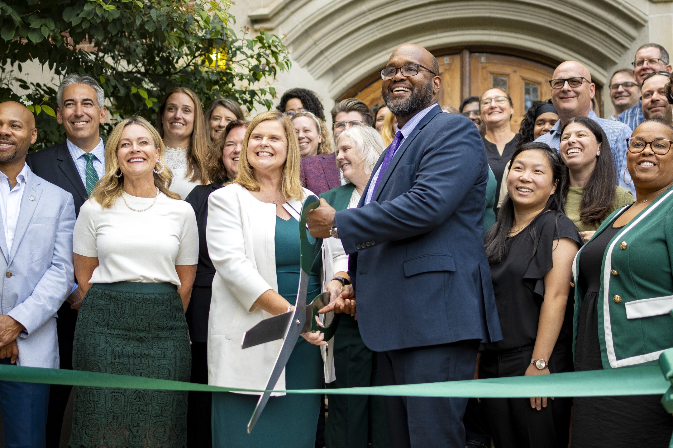 Image of MSU Honors College Interim Dean Glenn Chambers and Associate Vice President for Residential and Hospitality Services Patricia A.R. Martinez holding giant scissors to cut the ribbon at the entrance of Campbell Hall. They are surrounded by various MSU staff and leadership with enthusiastic expressions and business professional apparel.