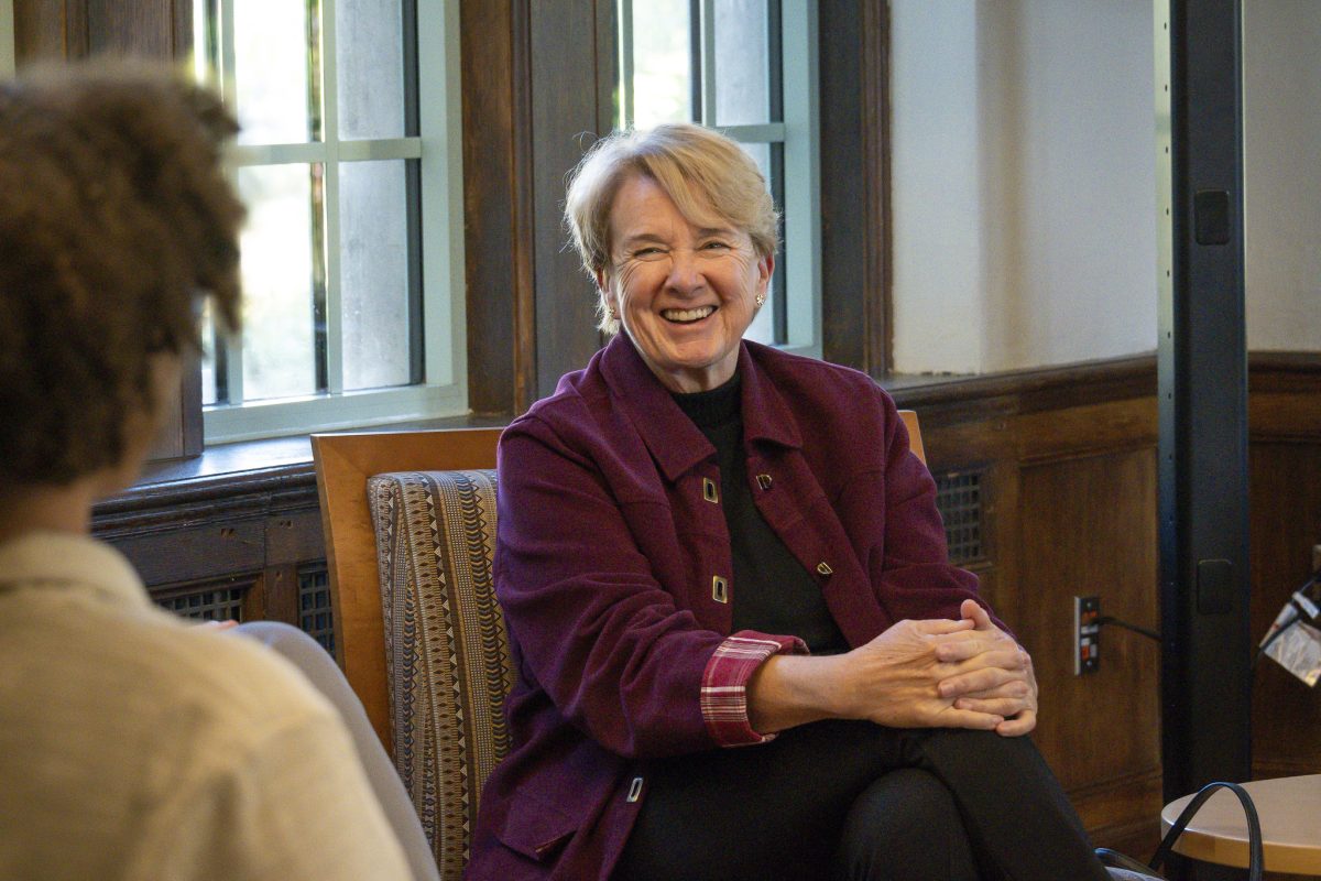 Image of Joanne Maguire smiling joyfully at a student inside Michigan State University's Campbell Hall. Maguire is in a dark purple jacket in black pants with hands clasped over one knee. Rows of windows are in the background.