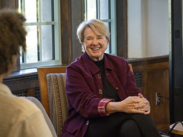 Image of Joanne Maguire smiling joyfully at a student inside Michigan State University's Campbell Hall. Maguire is in a dark purple jacket in black pants with hands clasped over one knee. Rows of windows are in the background.