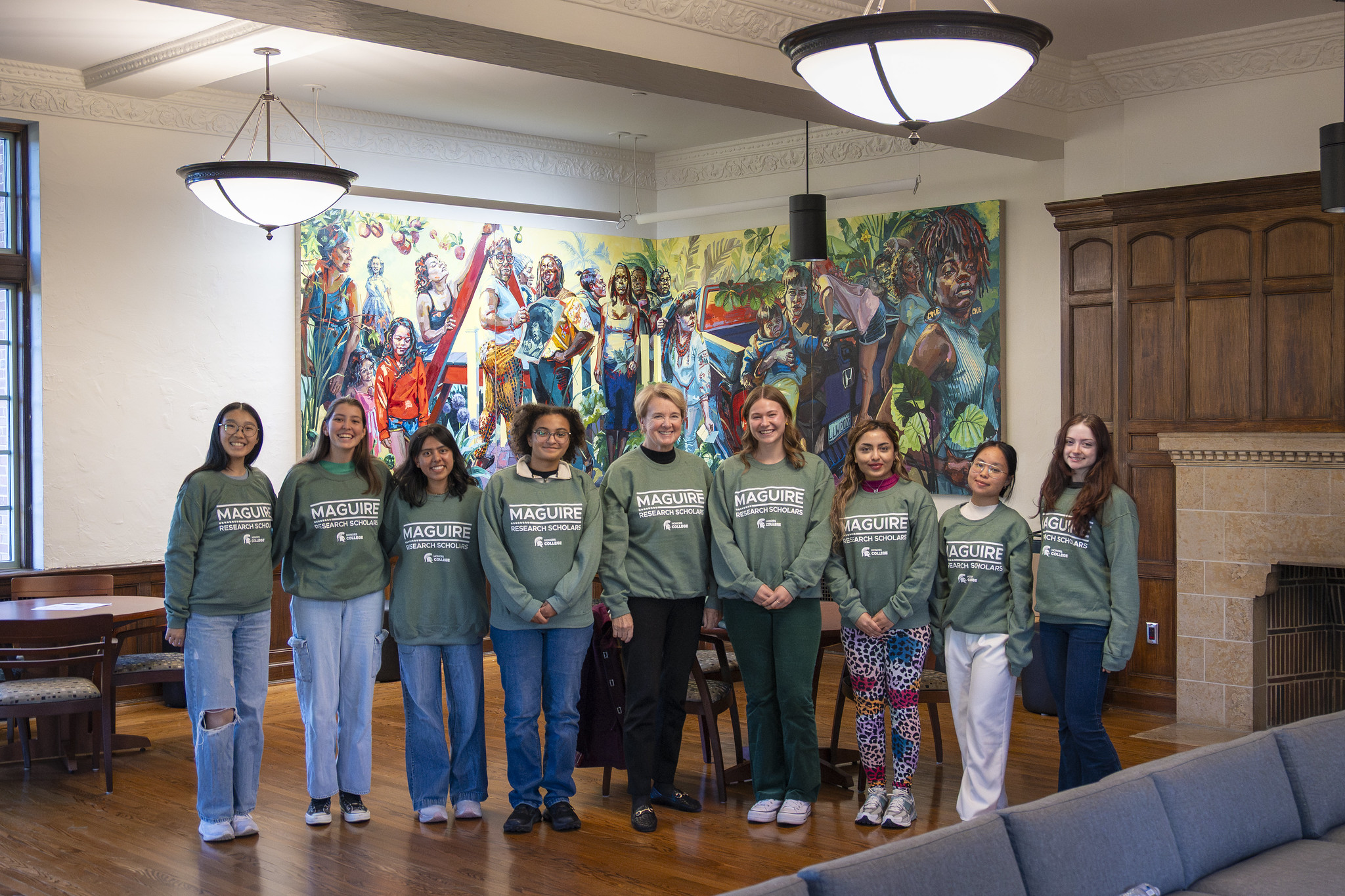 Image of alumna Joanne Maguire and eight Honors College Research Scholars, standing in a line and smiling at the camera. All of them are in green shirts with white text that says "Maguire Research Scholars. Honors College". A colorful mural is in the background.