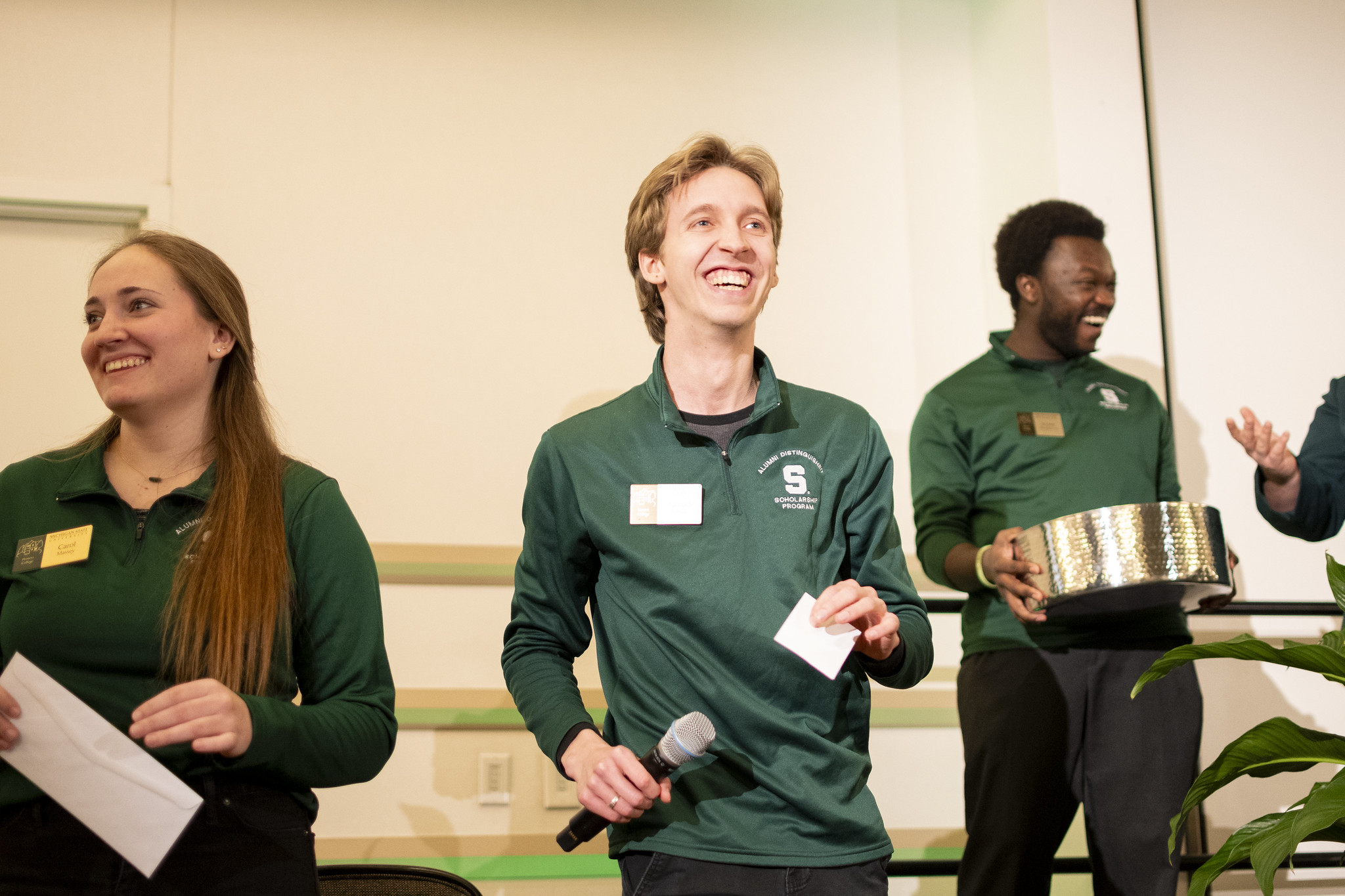 H-STAR leaders Carol Massey, Samuel Rabick, and Jerome Hamilton Jr. standing and smiling in different directions. Massey is looking to the right, holding a white envelope. Rabick is in the middle holding a microphone. Hamilton is looking off to the left while holding a giant metal container. All three are in dark green quarter-zips with white embroidery that reads: Alumni Distinguished Scholarship Program.