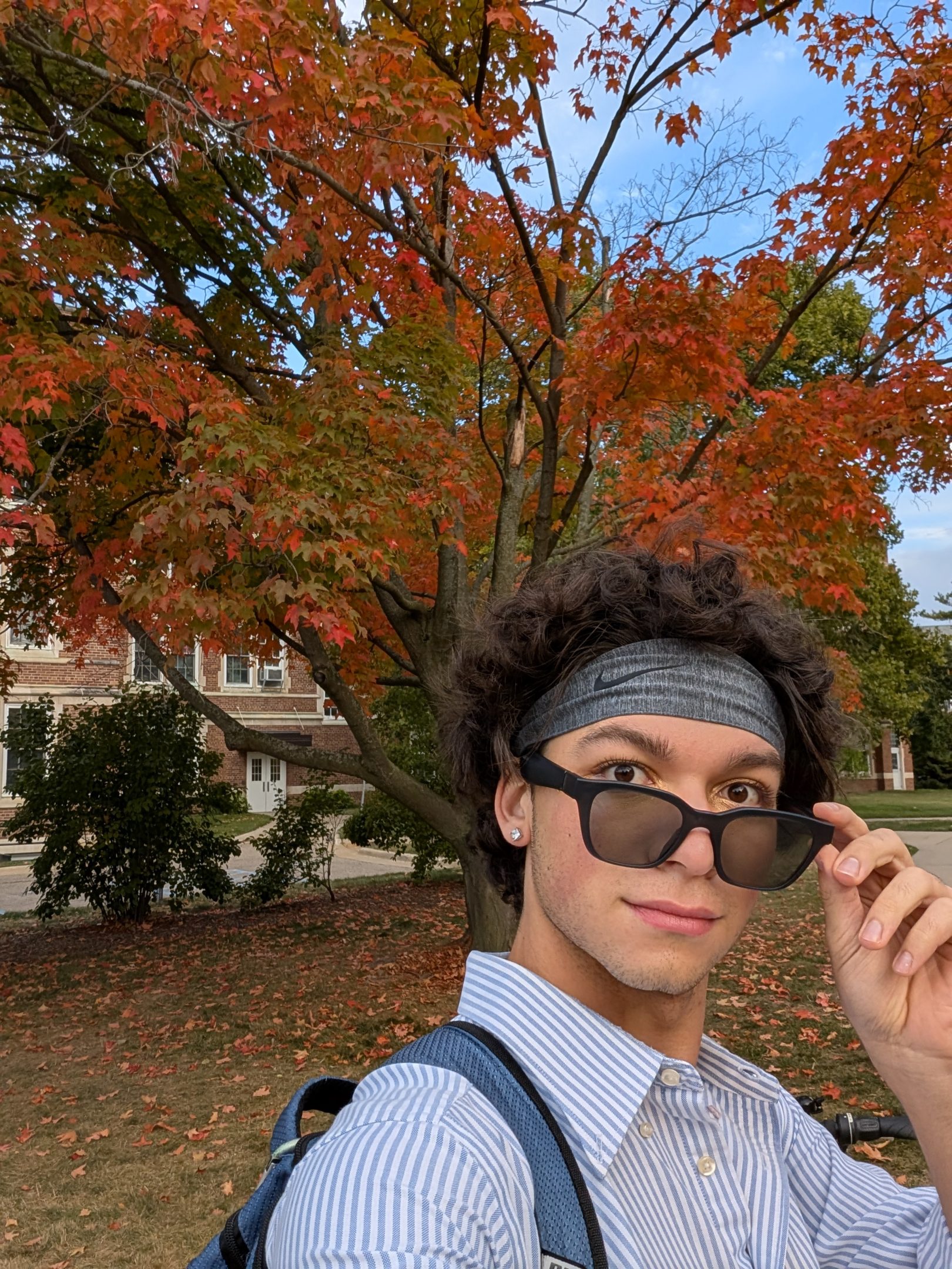 Image of H-STAR leader and Michigan State University student Camden Kimmel pulling down sunglasses and looking at the camera with a wide-eyed expression. Kimmel is in a light blue and white striped button-up shirt and grey bandana, with a backpack on. A tree full of orange and red leaves is in the background.