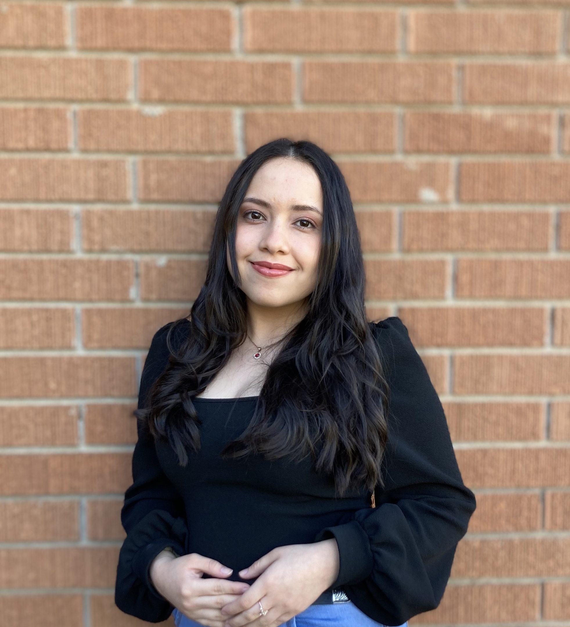 Image of H-STAR leader and current Michigan State University student Evelyn Gomez Recinos leaning against a brick wall and smiling softly. She is wearing a black top and blue jeans.